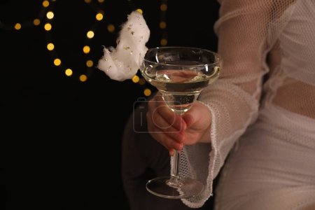 Woman holding glass of cocktail decorated with tasty cotton candy on black background with blurred lights, closeup. Space for text