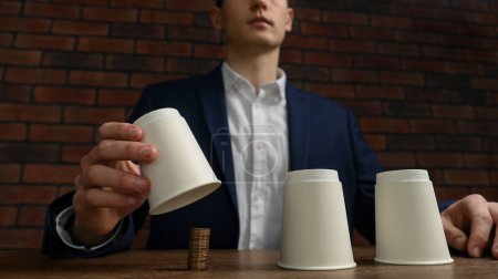Shell game. Man showing coins under cup at wooden table, low angle view