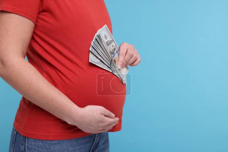 Téléchargez les photos : Mère porteuse. Femme enceinte avec des billets en dollars sur fond bleu clair, gros plan. Espace pour le texte - en image libre de droit