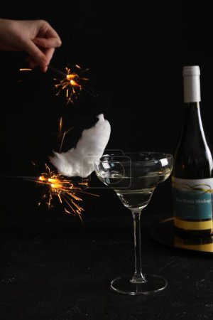 Cocktail with tasty cotton candy and bottle of alcohol drink on dark textured table. Woman holding sparkler against black background, closeup