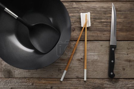 Photo for Black metal wok, chopsticks, knife and spatula on wooden table, flat lay - Royalty Free Image