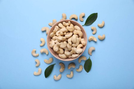 Many tasty cashew nuts and leaves on light blue background, top view