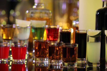 Different shooters in shot glasses and bottles on mirror surface against blurred background. Alcohol drink