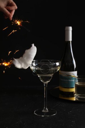 Cocktail with tasty cotton candy and bottle of alcohol drink on dark textured table. Woman holding sparkler against black background, closeup
