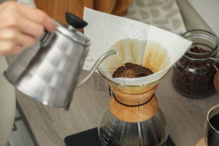 Woman pouring hot water into glass chemex coffeemaker with paper filter and coffee at countertop in kitchen, closeup