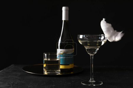Cocktail with tasty cotton candy and bottle of alcohol drink on dark textured table against black background