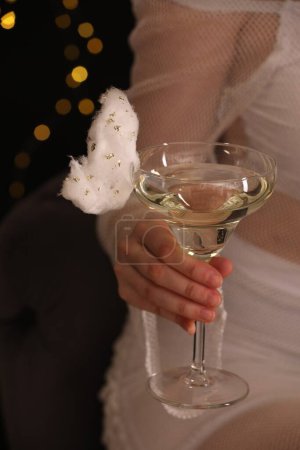Woman holding glass of cocktail decorated with tasty cotton candy on black background with blurred lights, closeup