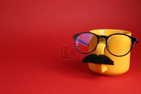 Téléchargez les photos : Visage d'homme en tasse, fausse moustache et lunettes sur fond rouge. Espace pour le texte - en image libre de droit