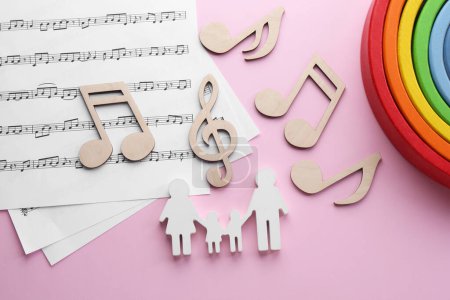 Babylieder. Notenblätter, Familienfiguren, Holznotizen und Spielzeugregenbogen auf rosa Hintergrund, flache Lage