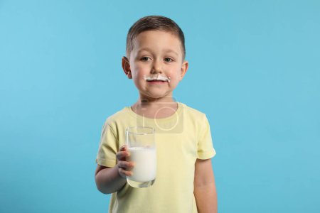 Téléchargez les photos : Garçon mignon avec moustache au lait tenant un verre de boisson laitière savoureuse sur fond bleu clair - en image libre de droit