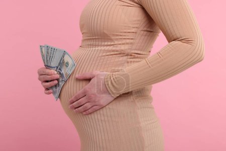 Téléchargez les photos : Mère porteuse. Femme enceinte avec des billets en dollars sur fond rose, gros plan - en image libre de droit