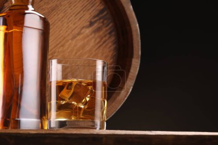 Whisky con cubitos de hielo en vidrio, botella y barril sobre mesa de madera sobre fondo negro, espacio para texto