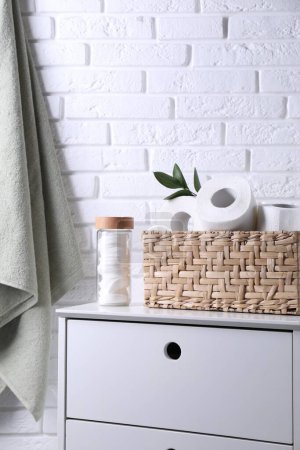 Toilet paper rolls in wicker basket, floral decor and cotton pads on chest of drawers