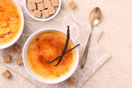 Deliciosa crema brulee en cuencos, vainas de vainilla, cubos de azúcar y cuchara en la mesa de textura ligera, vista superior