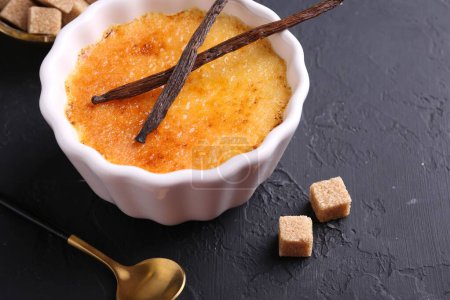 Photo for Delicious creme brulee in bowl, vanilla pods, sugar cubes and spoon on dark gray textured table, closeup - Royalty Free Image
