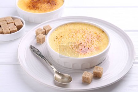 Téléchargez les photos : Délicieuse crème brulée dans des bols, des cubes de sucre et une cuillère sur une table en bois blanc - en image libre de droit