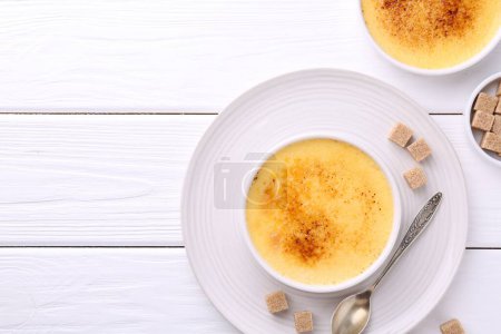 Foto de Deliciosa crema brulee en cuencos, terrones de azúcar y cuchara en la mesa de madera blanca, la puesta plana. Espacio para texto - Imagen libre de derechos