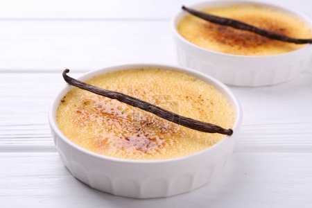 Téléchargez les photos : Délicieuse crème brulée dans des bols et gousses de vanille sur table en bois blanc, gros plan - en image libre de droit