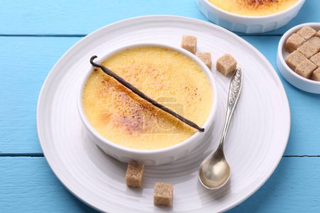Téléchargez les photos : Délicieuse crème brulée dans un bol, gousse de vanille, cubes de sucre et cuillère sur table en bois bleu clair - en image libre de droit