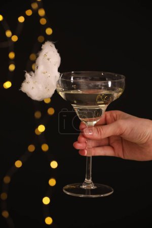 Woman holding glass of cocktail decorated with tasty cotton candy on black background with blurred lights, closeup