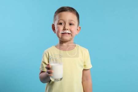 Téléchargez les photos : Garçon mignon avec moustache au lait tenant un verre de boisson laitière savoureuse sur fond bleu clair - en image libre de droit