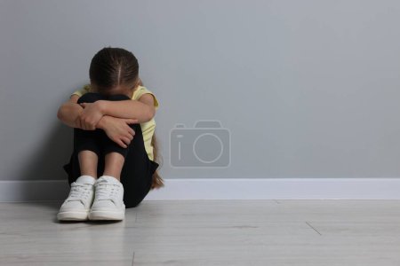 Abuso infantil. Muchacha molesta sentada en el suelo cerca de la pared gris, espacio para el texto