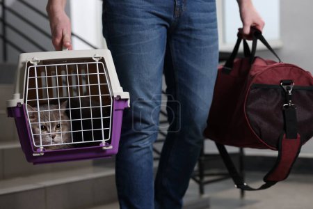 Travel with pet. Man holding carrier with cute cat and bag indoors, closeup