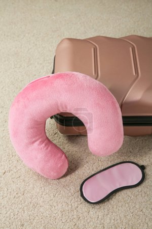Pink travel pillow, suitcase and sleep mask on beige rug