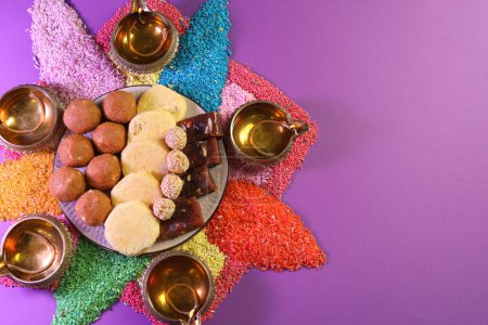 Diwali-Feier. Leckere indische Süßigkeiten, Diya-Lampen und bunte Rangoli auf violettem Tisch, flach gelegt. Raum für Text