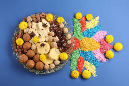 Diwali celebration. Tasty Indian sweets, colorful rangoli and chrysanthemum flowers on blue table, flat lay