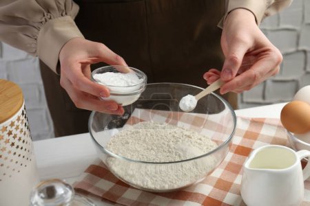 Woman with spoon and bowl of baking powder at white table, closeup
