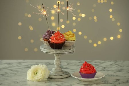 Different colorful cupcakes with sparklers on white marble table against blurred lights