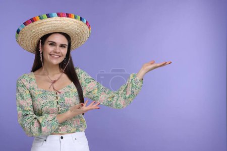 Mujer joven con sombrero mexicano mostrando algo sobre fondo violeta. Espacio para texto