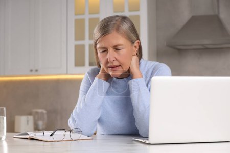 Menopause. Woman suffering from pain in neck at table in kitchen, space for text