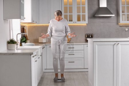 Foto de Menopausia, aumento de peso. Mujer preocupada de pie en balanzas de piso en la cocina - Imagen libre de derechos