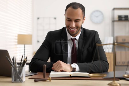 Photo for Smiling lawyer reading book at table in office - Royalty Free Image