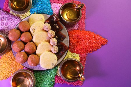 Diwali-Feier. Leckere indische Süßigkeiten, Diya-Lampen und bunte Rangoli auf violettem Tisch, flach gelegt