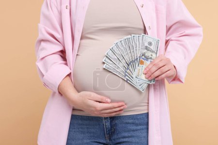 Téléchargez les photos : Mère porteuse. Femme enceinte avec des billets en dollars sur fond beige, gros plan - en image libre de droit