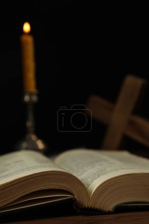 Kerze, Bibel und Kreuz auf Holztisch vor schwarzem Hintergrund