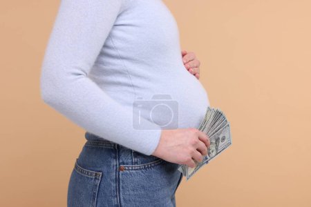 Téléchargez les photos : Mère porteuse. Femme enceinte avec des billets en dollars sur fond beige, gros plan - en image libre de droit