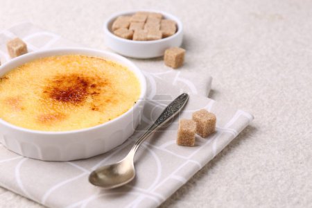 Foto de Deliciosa crema brulee en tazón, terrones de azúcar y cuchara en mesa de textura ligera, primer plano - Imagen libre de derechos