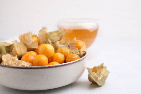 Ripe physalis fruits with calyxes in bowl on white tiled table, closeup. Space for text