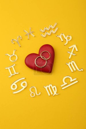 Foto de Signos del zodíaco, corazón rojo y anillos de boda sobre fondo amarillo, posición plana - Imagen libre de derechos
