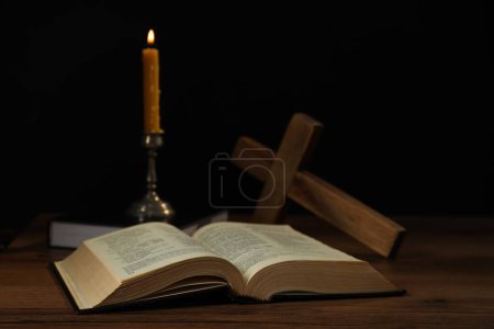 Church candle, Bible and cross on wooden table against black background