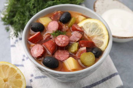 Meat solyanka soup with thin dry smoked sausages in bowl on grey table, closeup