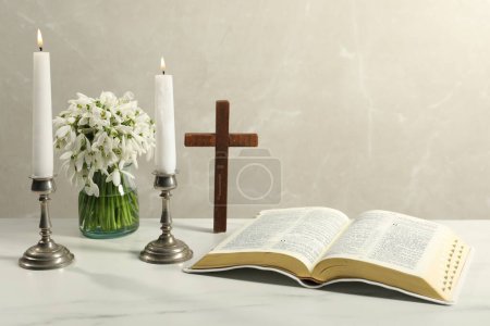 Burning church candles, Bible, wooden cross and flowers on white marble table