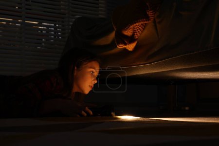 Téléchargez les photos : Petite fille avec lampe de poche à la recherche de monstre sous le lit la nuit - en image libre de droit