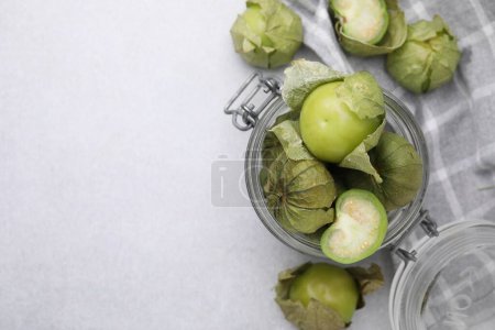 Fresh green tomatillos with husk in glass jar on light table, flat lay. Space for text