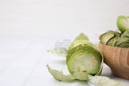 Fresh green tomatillos with husk in bowl on white tiled table, closeup. Space for text
