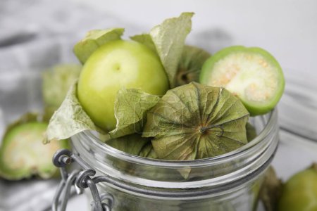 Fresh green tomatillos with husk in glass jar on table, closeup